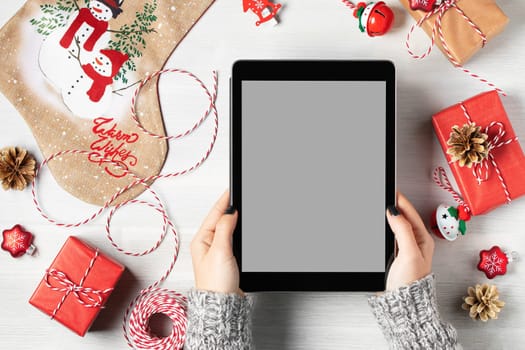 Tablet with a blank screen in a woman's hands surrounded by Christmas decorations and gifts, mockup for advertising.