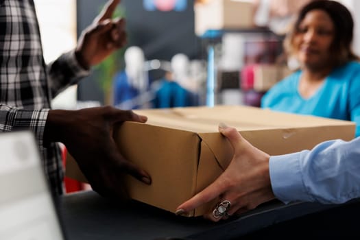 Shopaholic man holding cardboard box at counter desk, after buying fashionable merchandise. African american man shopping for stylish clothes, discussing about material in modern boutique