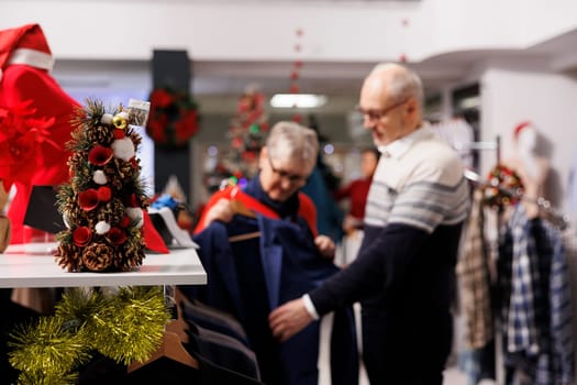 Selective focus of customers reviewing suit jacket on racks, looking for festive clothing to wear at christmas eve dinner. Persons checking blazers fabric in retail store, formal attire outlet.