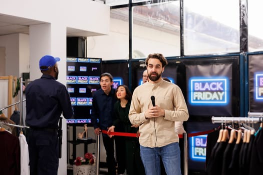 Young man TV reporter covering news real-time, live streaming during Black Friday sales. Correspondent standing at store entrance talking into microphone making live newscast about shopping madness