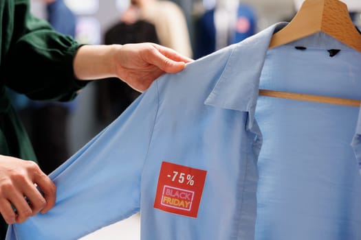 Female customer looking at shirt with red price tag while shopping on Black Friday in fashion mall. People buy clothes at discounted prices, make purchases during seasonal big sales in clothing store