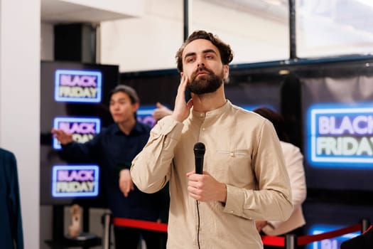 TV reporter live broadcasting talking about Black Friday lines in local shopping mall, holding microphone and looking at camera. Young man correspondent covering news about sales season in retail