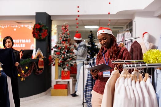 African american retail employee looking at list of merchandise in clothing store, working on stock inventory during sales season. Woman manager checking racks of trendy clothes.