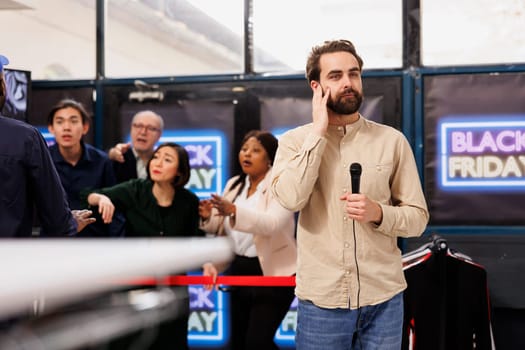 Young man TV news reporter standing with microphone against angry crazy crowd of shoppers waiting for discounts. Journalist correspondent making special reportage about Black Friday event