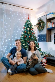 Happy parents with a little girl and a cat sit near the Christmas tree on the floor. High quality photo
