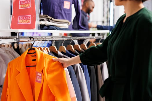 Customer examining clothes hanging on rack in clothing store, doing purchases at shopping mall during Black Friday sales. Woman looking at red price tag while buying formal wear in fashion boutique