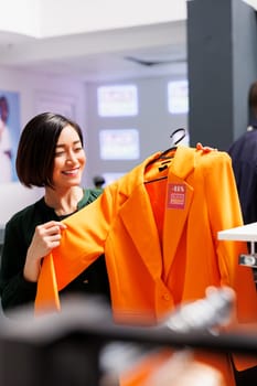 Black Friday. Happy young Asian woman customer standing in clothing store with trendy jacket in hands, enjoying spending money in fashion boutique during sales season. Happiness and shopping