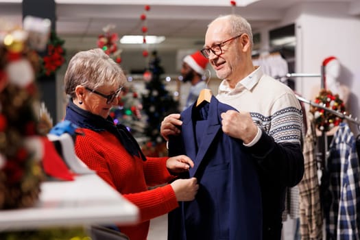 Elderly couple looking at blazers in retail store, searching for formal clothing to wear on christmas dinner celebration. Customers checking fabric of suit jacket for elegant outfit, festive decor.