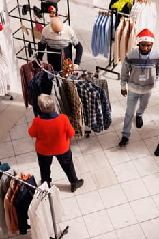 Various shoppers looking at clothing items on racks in shopping center while seeking for garments to purchase. Consumers going apparel shopping as gifts for festive season, Christmas eve.