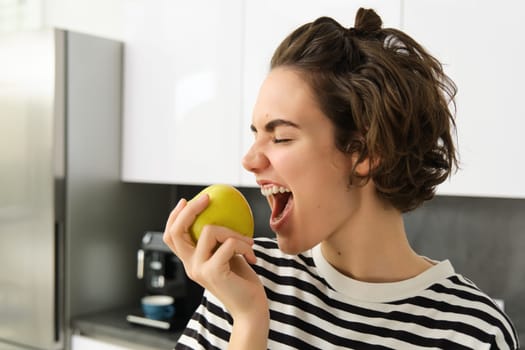 Close up portrait of smiling woman in the kitchen, holding an apple, eating fruit, having a healthy snack for lunch at home.