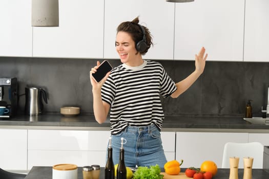 Portrait of happy dancing woman, cooking in the kitchen and listening music in headphones, chopping vegetables for salad and singing with carefree face.