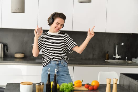 Dancing girl cooking salad in the kitchen, listening music in wireless headphones, holding smartphone in hand, making a meal, cooking at home.