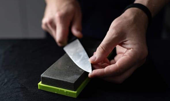 Man Uses Natural Sharpening Whetstone To Sharpen Big Kitchen Knife Against Black Background