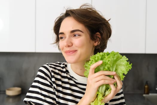 Portrait of young smiling woman, vegetarian washing lettuce leaves for salad, holding ingredients in hand, making a vegan meal, being on diet, standing in kitchen.