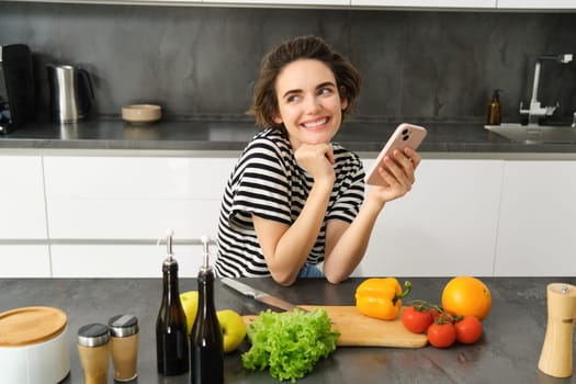 Cute young brunette woman cooking food in the kitchen, looking for healthy meal with vegetables online, using smartphone, sitting near counter and smiling.