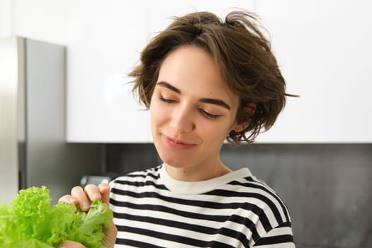 Close up portrait of smiling, beautiful female model, making a salad, holding green lettuce leaf, preparing vegetables, cooking in the kitchen, standing in striped t-shirt.