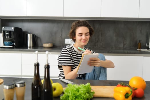 Smiling brunette woman in kitchen, writing down meal list for week, reading recipe, cooking salad, vegetable meal at home. Lifestyle concept