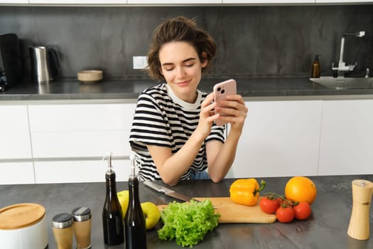 Portrait of young woman with smartphone, sitting in the kitchen and cooking salad, searching for healthy recipe online, has vegetables and chopping board on the table.
