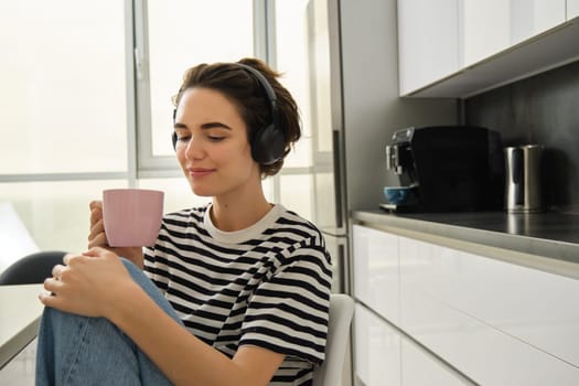Portrait of smiling, relaxed woman, drinks warm tea and listens music or e-book in kitchen. Lifestyle and people concept
