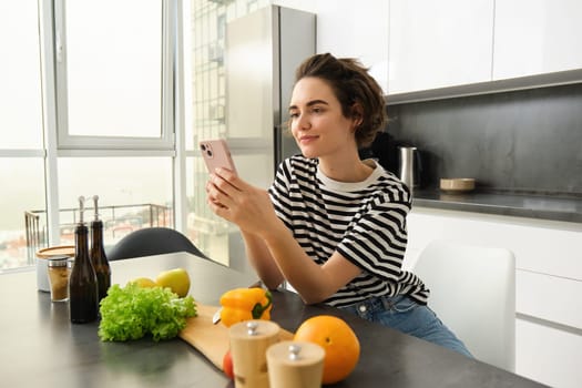 Portrait of brunette girl cooking food in the kitchen, searching recipes on social media app, holding mobile phone, standing near chopping board and vegetables, preparing healthy vegetarian meal.