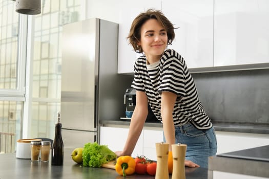 Portrait of happy, healthy young woman, vegetarian making herself salad, posing near vegetables on kitchen, chopping ingredients for vegan meal.
