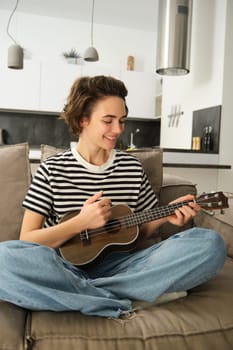 Vertical portrait of happy young woman playing ukulele, learns new musical instrument, sits on sofa at home, smiling with joy.