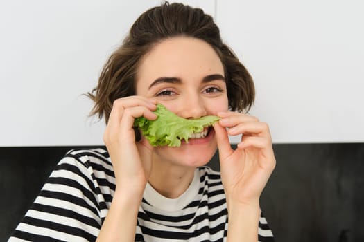 Close up portrait of young woman, vegetarian girl, likes eating vegetables, posing with lettuce leaf and smiling, posing in the kitchen. Concept of healthy food and diet.