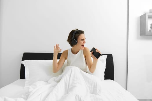 Carefree modern woman singing in her bedroom, into smartphone microphone, listening to music in wireless headphones. Lifestyle concept