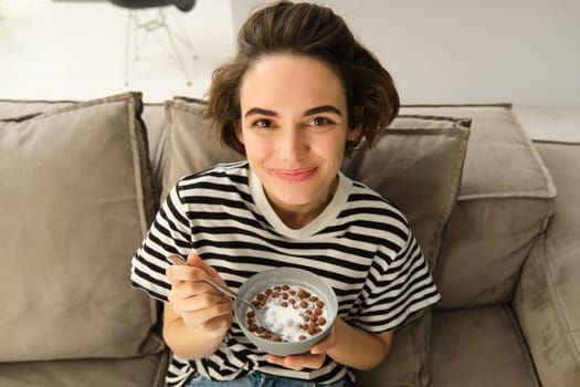 Close up of cute young female model, eating cereals with milk, enjoys her breakfast on sofa in living room, smiling and looking happy. People and food concept