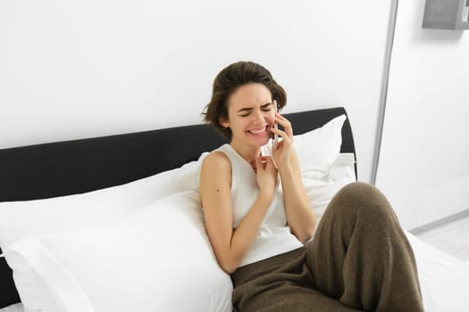 Beautiful, carefree modern woman, laughing over the phone, calling friend and chatting, resting in bed, spending time at home. Leisure and people concept.