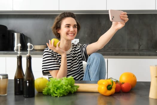 Portrait of beautiful smiling woman in the kitchen, taking selfie with apple, cooking healthy meal from vegetables, chopping food ingredients and recording video on mobile phone.