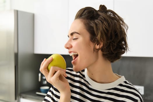 Close up portrait of young brunette woman biting an apple with pleasure, has pleased smile on her face, standing in the kitchen, having healthy snack for lunch, eating fruits.