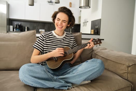 Young woman playing ukulele with passion, singing song, strumming strings, sitting on sofa in living room at home. Lifestyle and music concept