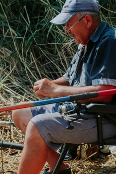 One Caucasian elderly happy man in old dirty clothes puts a worm on the hook of a fishing rod while sitting on a chair on the shore of a lake on a sunny summer day, side view close-up.