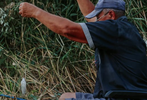 One Caucasian elderly man in old dirty clothes pulls out a fish caught with a fishing rod from the river while sitting on a chair on the shore of a lake on a sunny summer day, close-up side view.