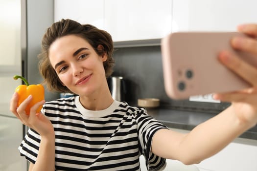 Cute and stylish lifestyle blogger, young woman taking selfie with fresh pepper, eating healthy vegetables, cooking salad in the kitchen and recording video on smartphone.