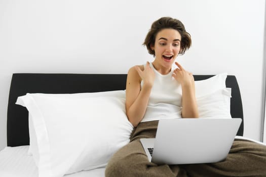 Portrait of woman sitting on bed with laptop, gasping and looking surprised at computer screen. Technology and lifestyle concept