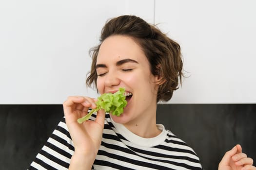 Close up of funny cute woman, vegetarian eating lettuce leaf and smiling, concept of healthy diet, girl likes vegetables, standing in the kitchen.