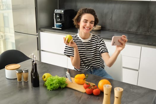 Portrait of beautiful young woman, food blogger recording video of her cooking in the kitchen, showing meal ingredients, preparing a meal on camera, using smartphone for live streaming.
