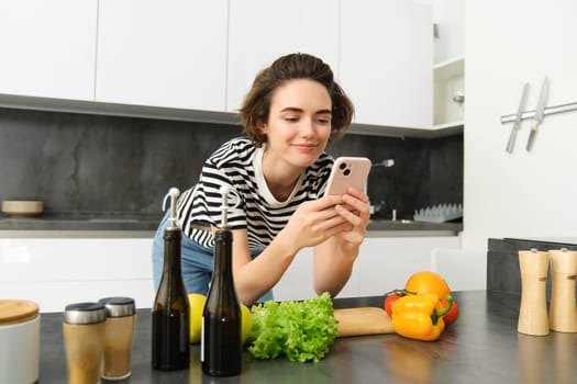Woman bending on kitchen counter with smartphone, looking for healthy recipe online on mobile phone, standing next to chopping board and vegetables, cooking ingredients.