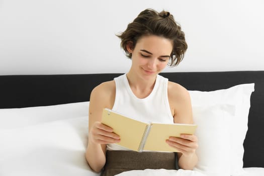 Portrait of woman reading her planner and starting the day, looking at notes, sitting on bed in morning.