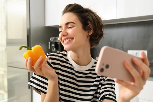 Smiling modern woman, vegetarian food blogger, taking selfie with fresh yellow sweet pepper, holding smartphone, recording a video blog for her social media account, posing in the kitchen.