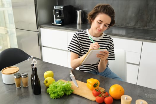 Young brunette woman writing down recipe, making notes in notebook while cooking a salad, thinking of grocery list, sitting in the kitchen with vegetables on the table.