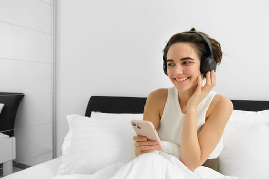 Beautiful girl in bed, listens to music in wireless headphones, using smartphone and smiling. Copy space