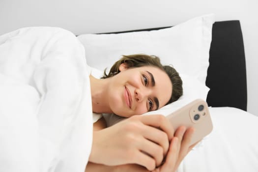 Close up portrait of lovely young woman in bed, lying and going to sleep, checking her phone messages, using smartphone and smiling.