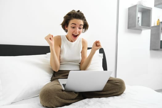 Portrait of woman sitting on bed, looking at laptop with excited, amazed face, celebrating, making fist pump, triumphing after winning on computer.