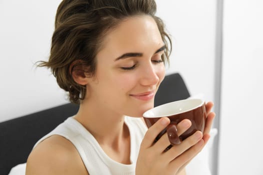 Close up portrait of brunette, smiling woman, smelling her coffee in cup, waking up and drinking cappuccino in bed.
