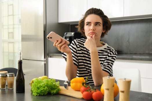 Portrait of young woman standing in the kitchen with vegetables and chopping board, holding smartphone, searching healthy diet recipes on mobile phone browser, cooking at home.