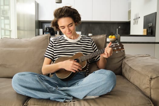 Lifestyle and hobbies concept. Young smiling woman on sofa, playing ukulele, singing and learning strumming pattern for favourite song.