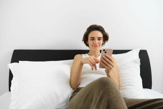 Portrait of young modern woman lying in her bed and using mobile phone, resting in bedroom and looking at smartphone screen.